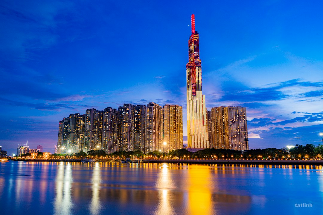 A tall illuminated skyscraper rising above apartment towers along a river at blue hour